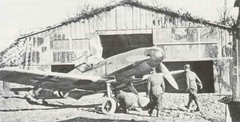 Bf 109 F steht vor einem als Scheune getarnten Hangar